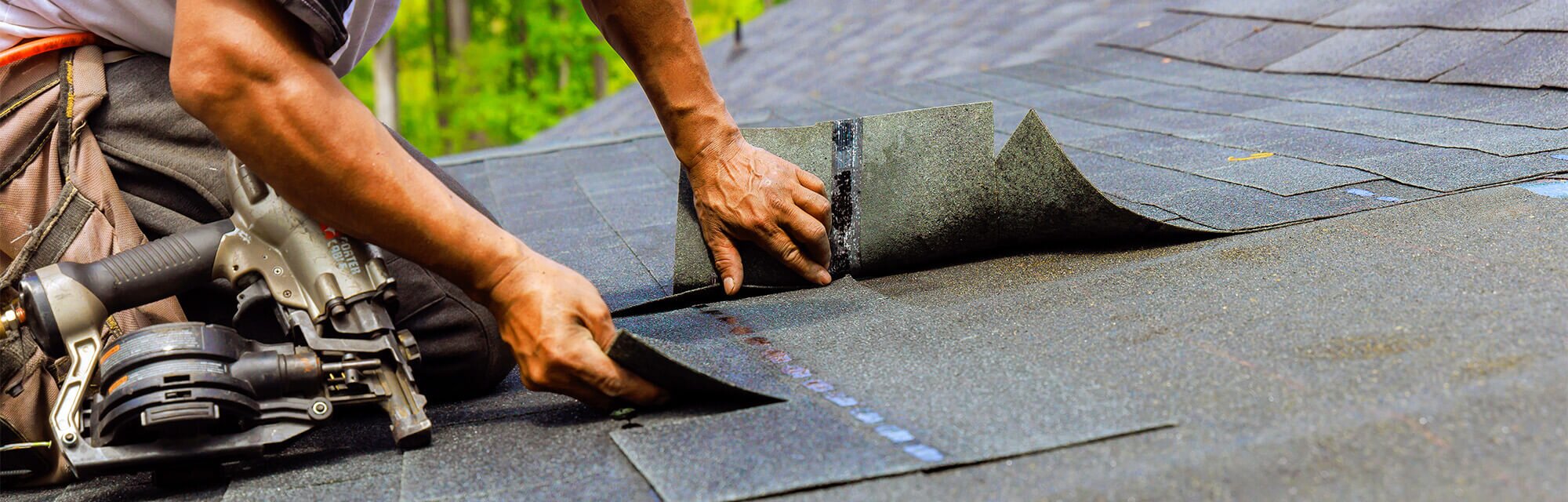 A person installs new black shingles on a roof, with a nail gun and nails nearby.