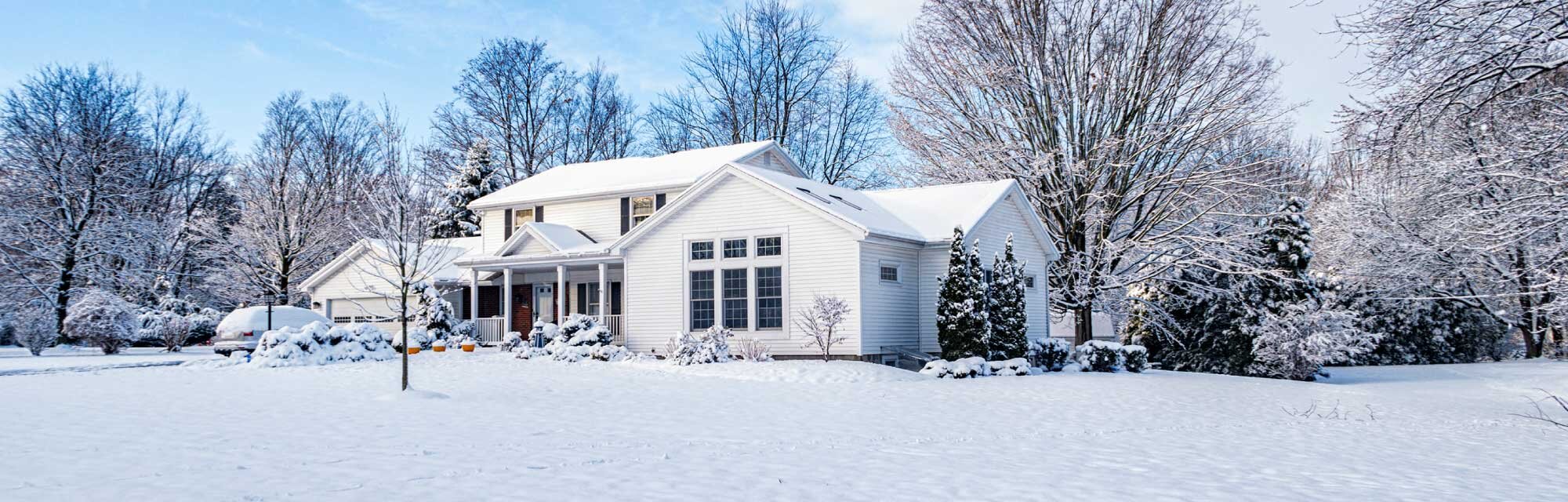 A white house is surrounded by trees with snow covering the roof and landscaping.