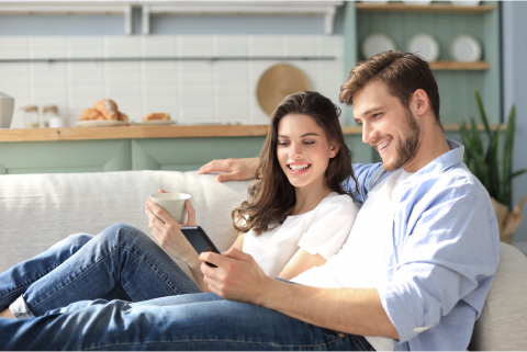 A man and woman sitting on a couch, both browsing the internet on mobile phone hold by men.