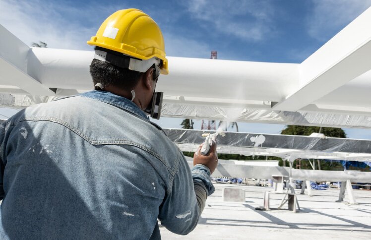 Worker in a yellow hard hat and denim jacket sprays a white metal structure outdoors under a bright blue sky.