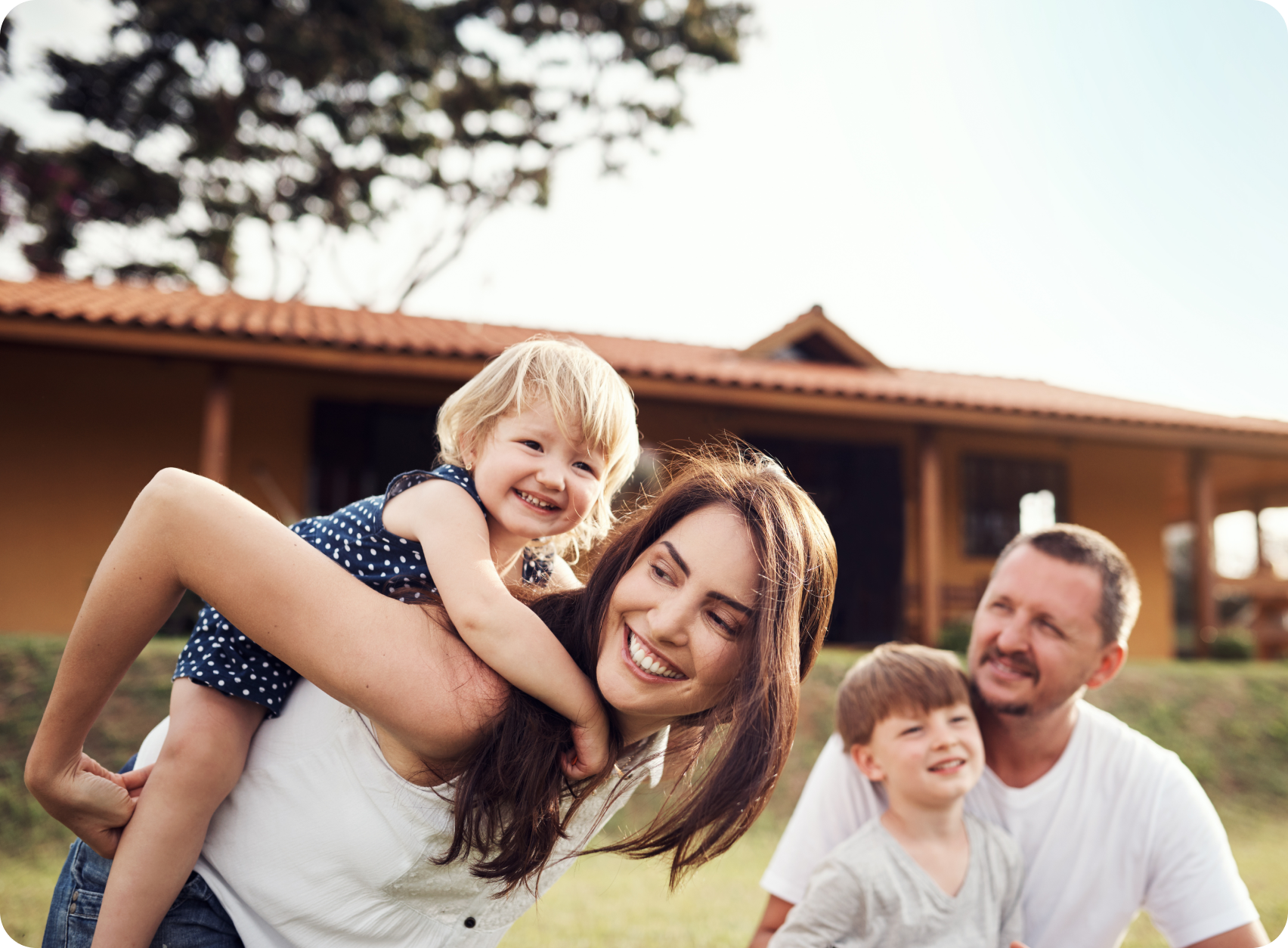 A family of four laughing together outdoors in front of a house, with a mother holding a young child on her back and a father sitting beside another child.
