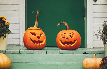 Two Halloween jack-o-lanterns rest on a home’s green porch with potted flowers to the left and the right. 