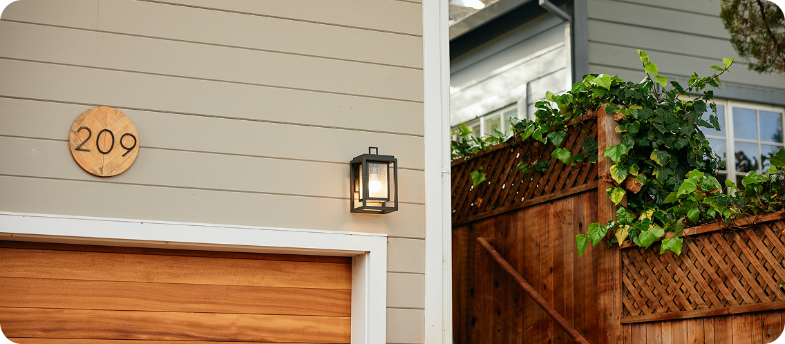 The exterior of a house showing a garage door, house number, and a wall lantern with greenery.