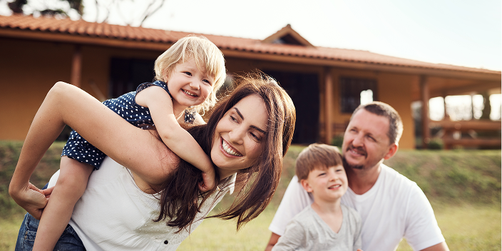 A family of four laughing together outdoors in front of a house, with a mother holding a young child on her back and a father sitting beside another child.

