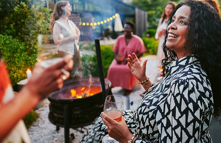 Woman with curly hair laughing joyfully at a backyard gathering with friends in a garden setting with string lights.