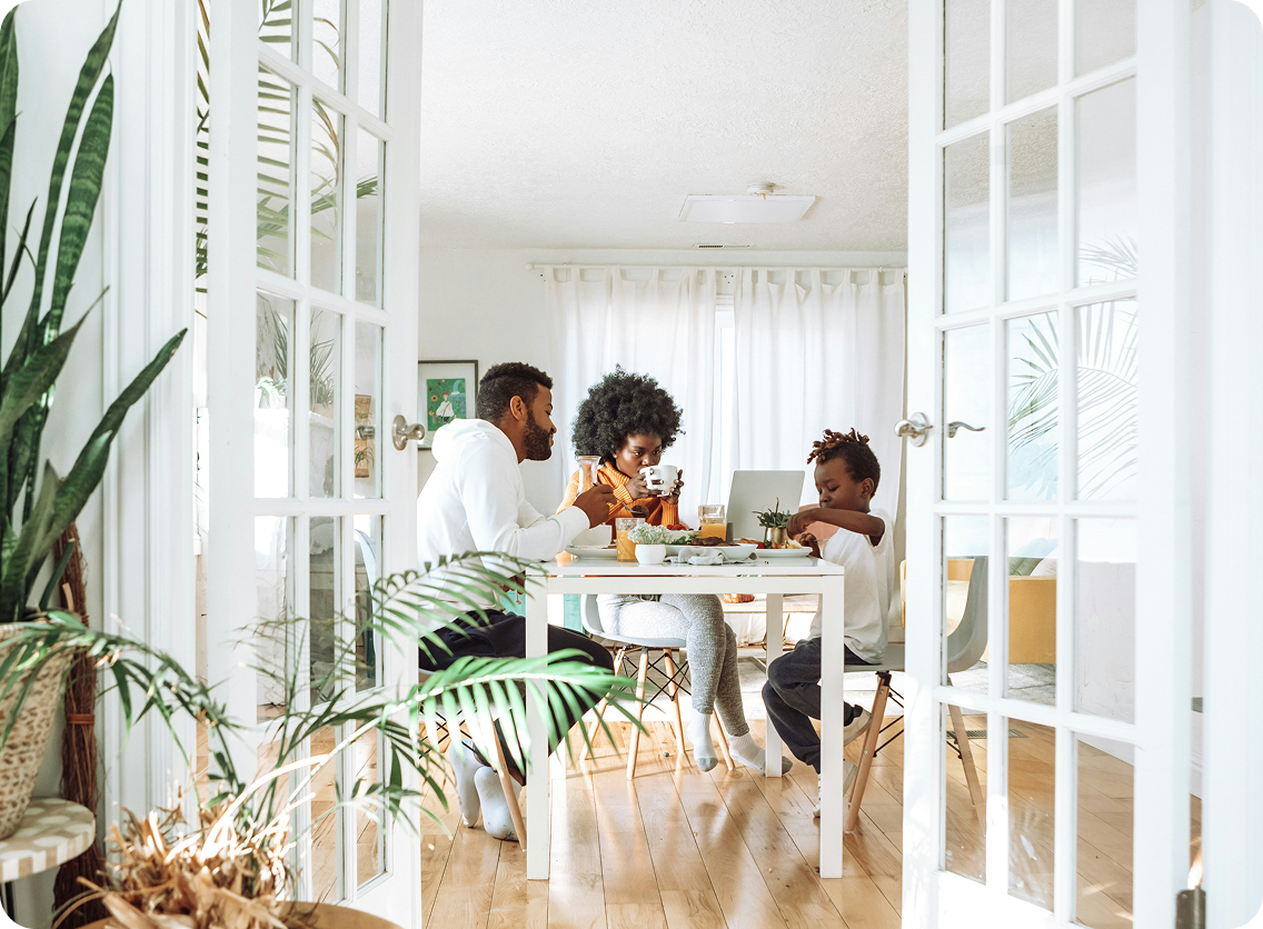 A family sitting together at a dining table in a bright, plant-filled home, viewed through open French doors.