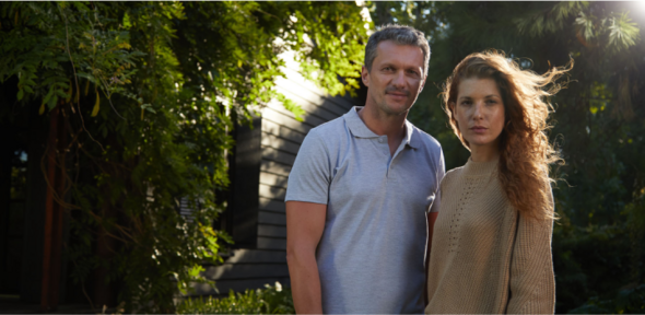 A couple standing in front of their house, surrounded by the lush green plants and trees of their garden.