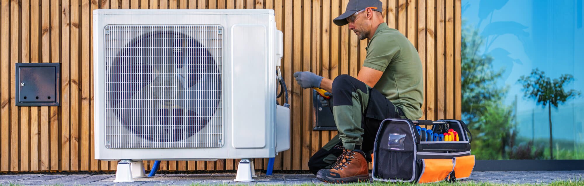 A repair professional wearing a cap and work gloves kneels next to an HVAC unit, with a toolbox nearby.