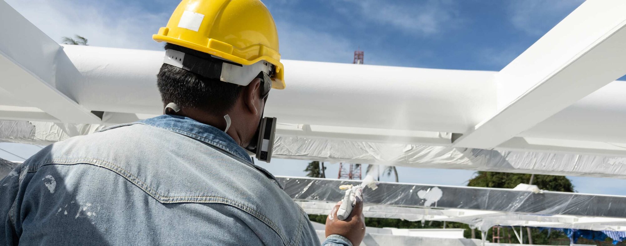 Worker in a yellow hard hat and denim jacket sprays a white metal structure outdoors under a bright blue sky.