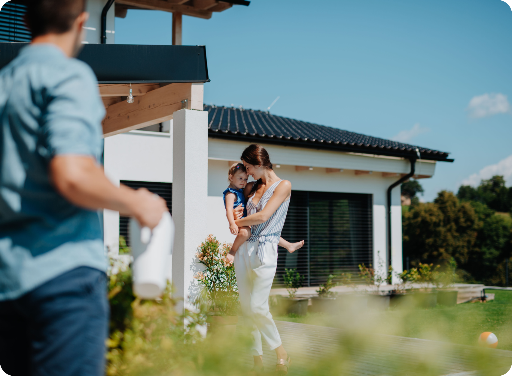A man in the foreground watering plants. A woman is holding a child outside a modern home.