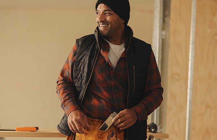 A homebuilder wearing a toolbelt stands smiling in a new construction home.