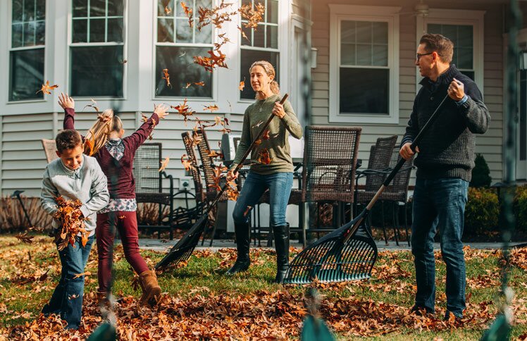 Two adult homeowners rake leaves in front of their home with two children.