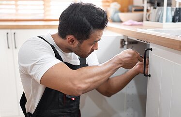 A homeowner wearing overalls performs maintenance on a kitchen sink while sun shines through the window blinds.