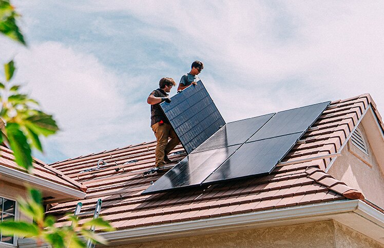 Two people stand on the roof of a home, installing solar panels on a sunny day.