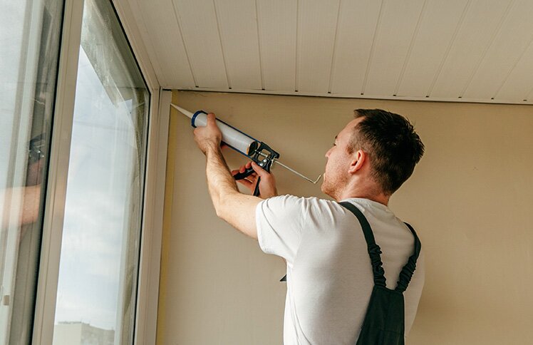 A homeowner re-applies caulk around the windows in his enclosed porch.