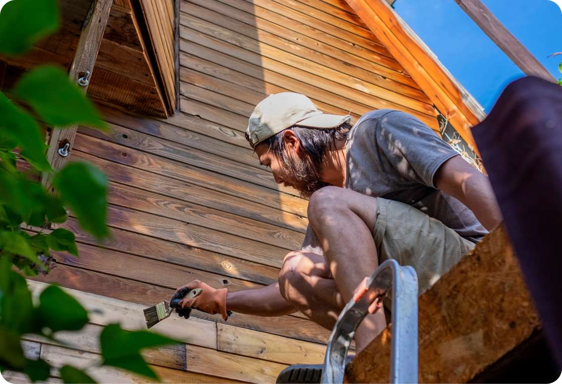 A man crouching down to stain his home's wood siding.