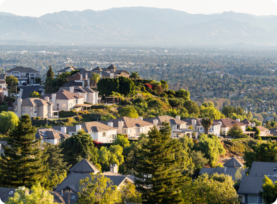 An aerial view of a California residential neighborhood with tree-lined streets, hillside homes, and mountains visible in the distance.