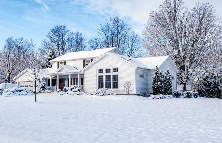 A white house is surrounded by trees with snow covering the roof and landscaping