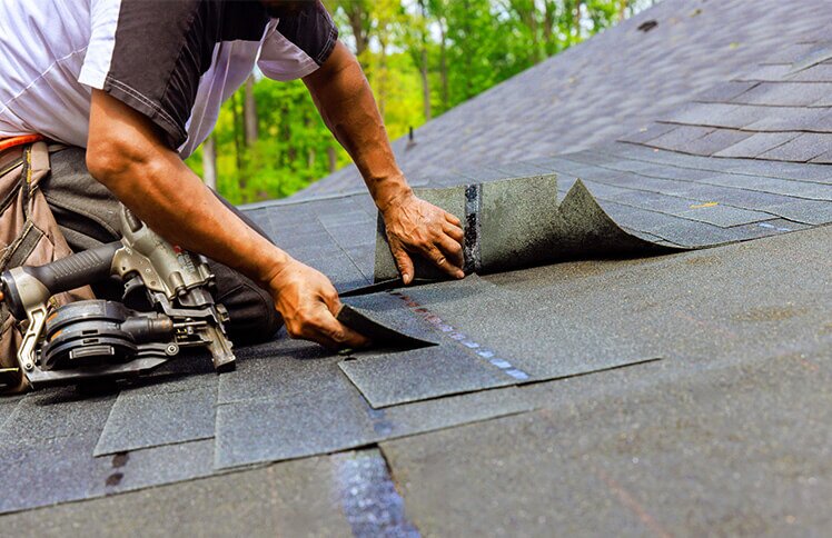 A person installs new black shingles on a roof, with a nail gun and nails nearby.