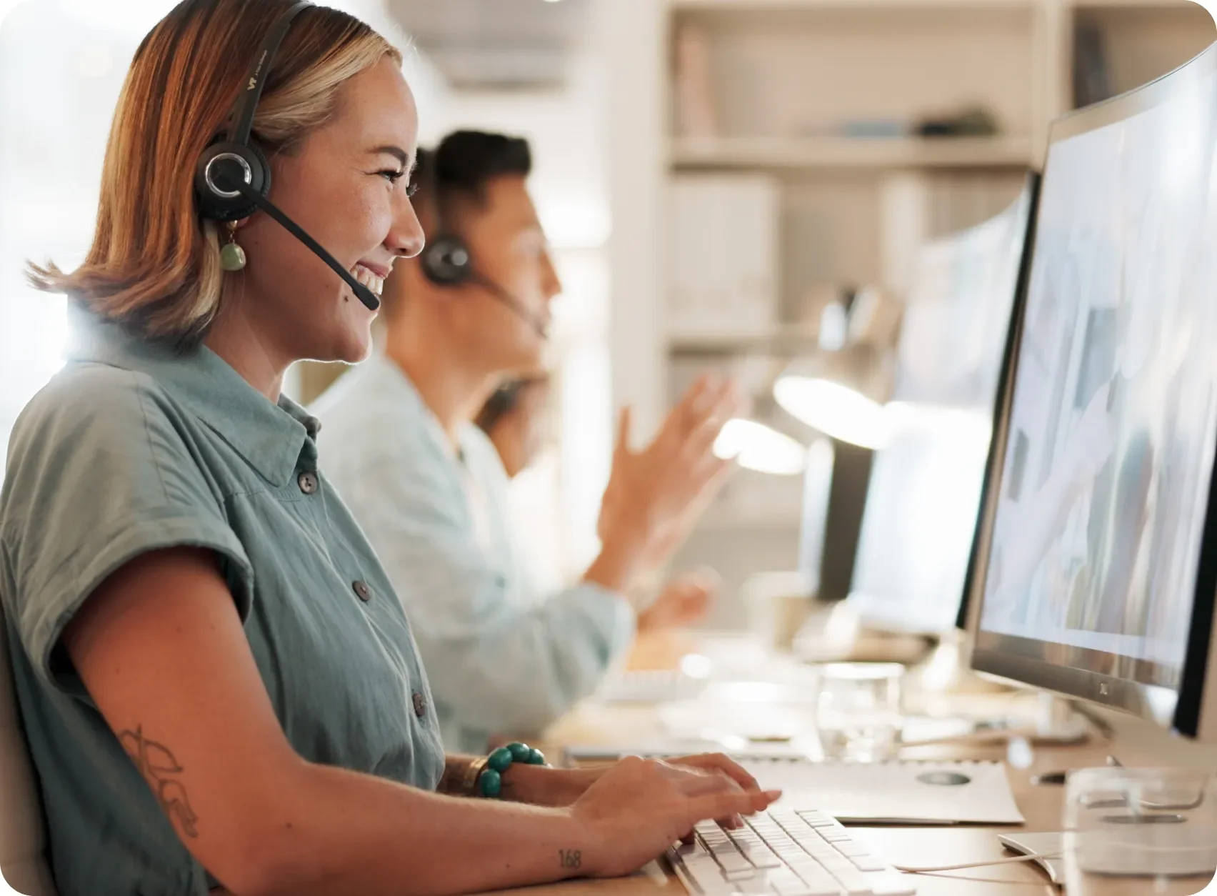 A woman wearing a headset smiles while working at a computer, with a colleague visible in the background.