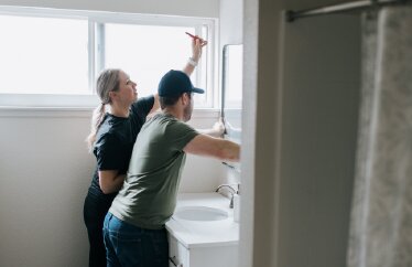 Two homeowners work together to hang a mirror above a sink in a bathroom.