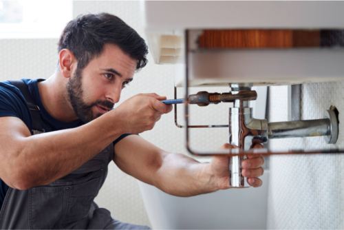 A plumber crouched under a toilet sink, using a wrench to fix a leaking pipe.