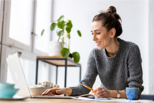 A woman sitting at a desk in a home office, smiling as she types on her laptop.