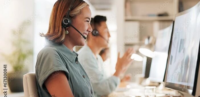 A woman wearing a headset smiles while working at a computer, with a colleague visible in the background.
