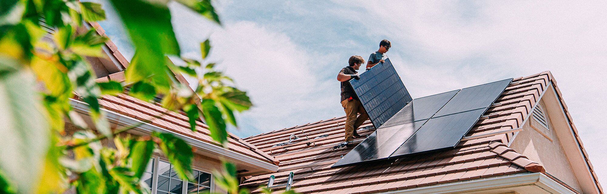 Two people stand on the roof of a home, installing solar panels on a sunny day.