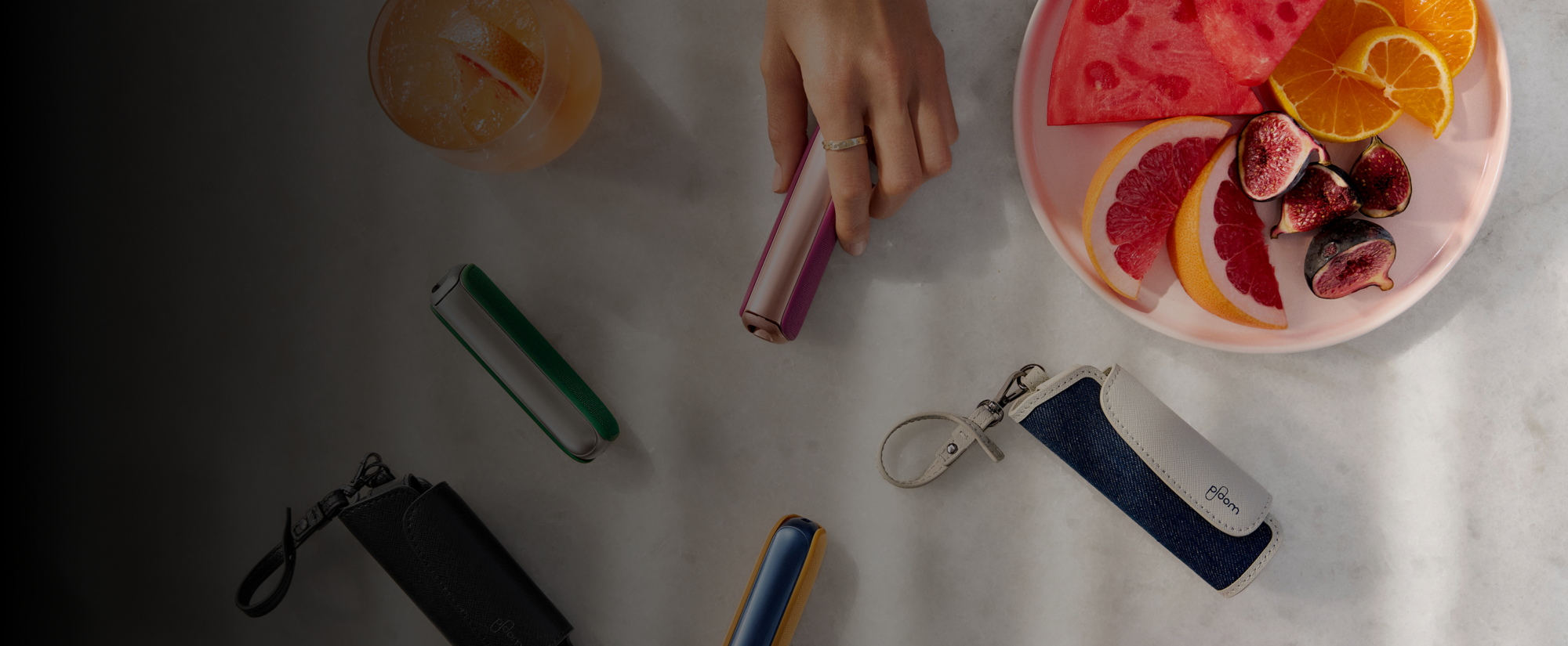 Hand taking a Ploom AURA Rose Gold device from a table, with Solo carry case and other accessories.