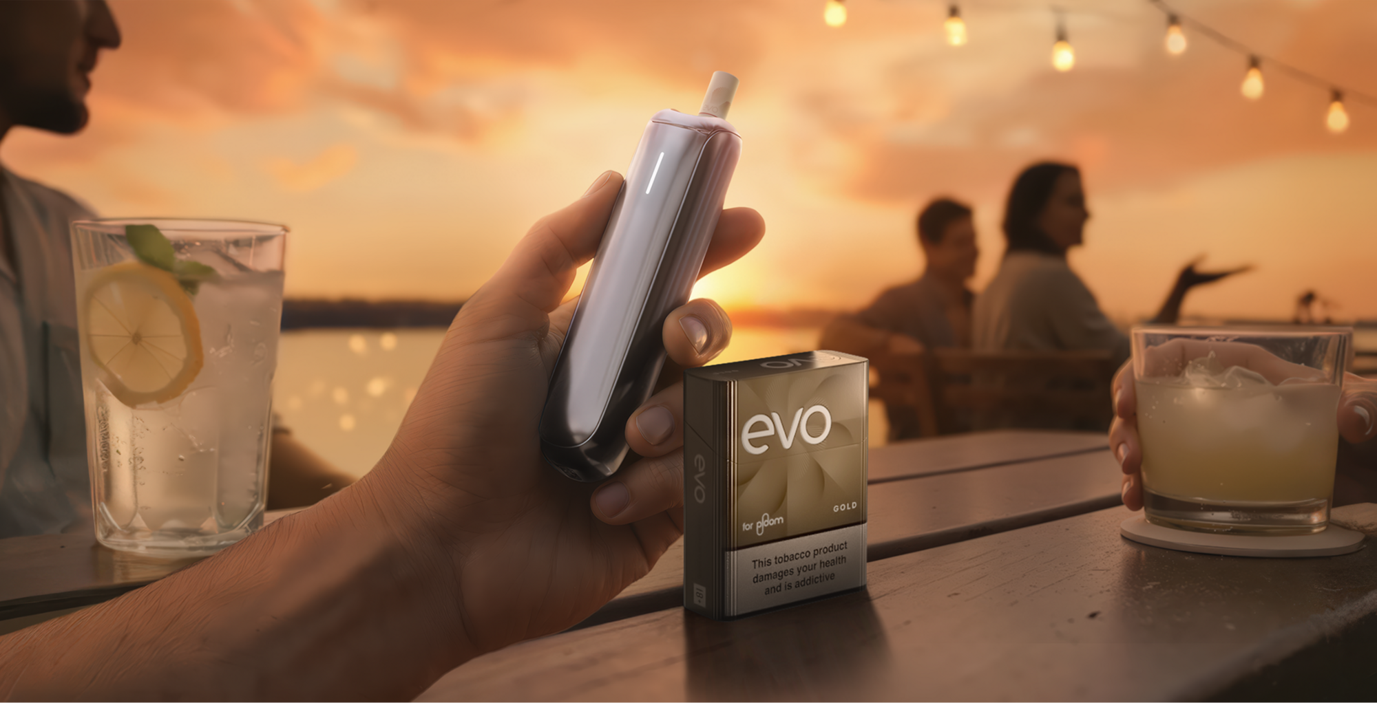 Person holding a Ploom device with a pack of EVO sticks at an outdoor table by the water at sunset