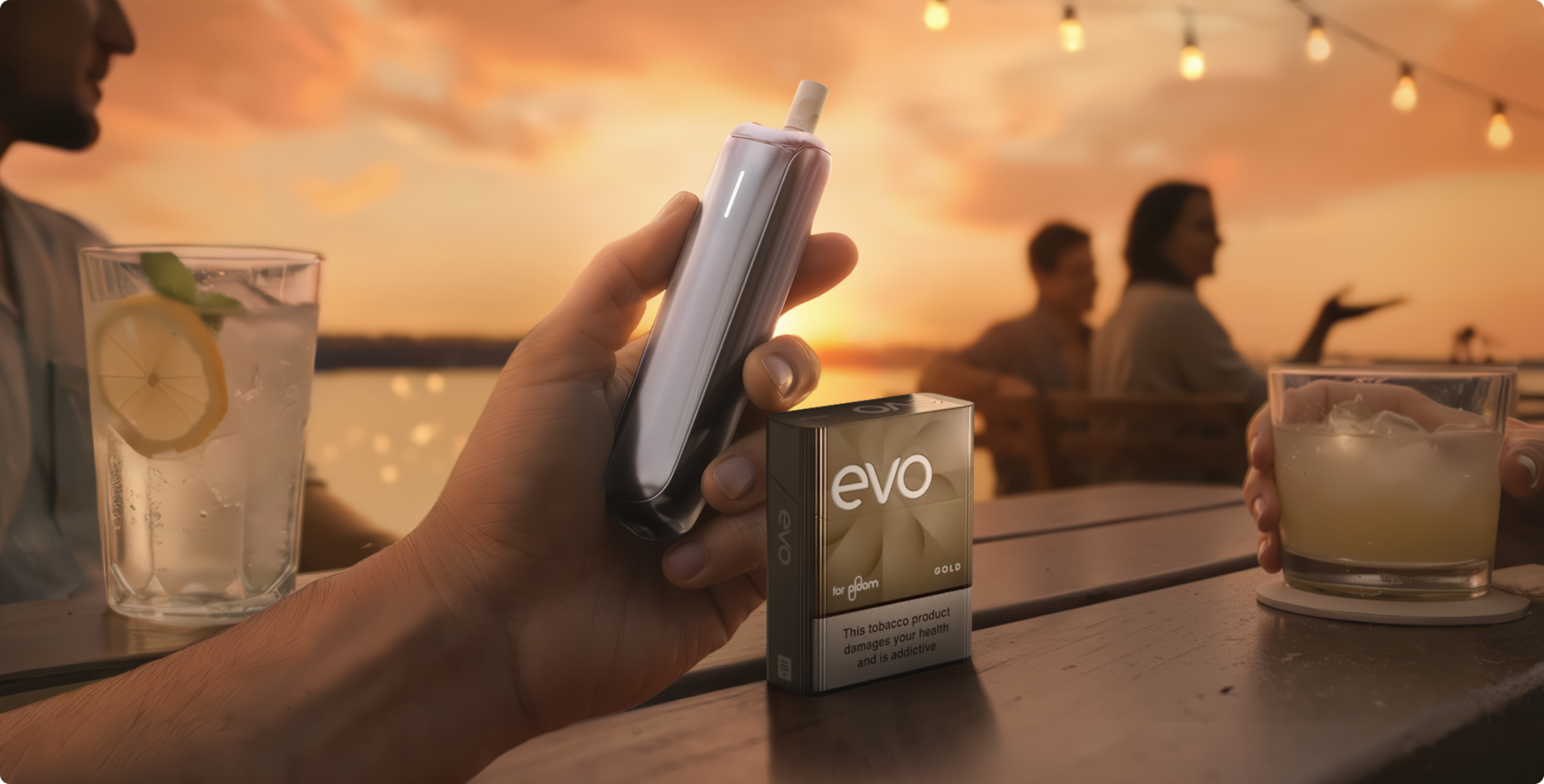 Person holding a Ploom device with a pack of EVO sticks at an outdoor table by the water at sunset