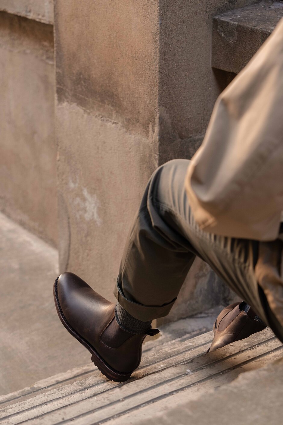 Person in olive pants wearing brown waxy calf leather Chelsea boots, sitting on concrete steps outdoors.