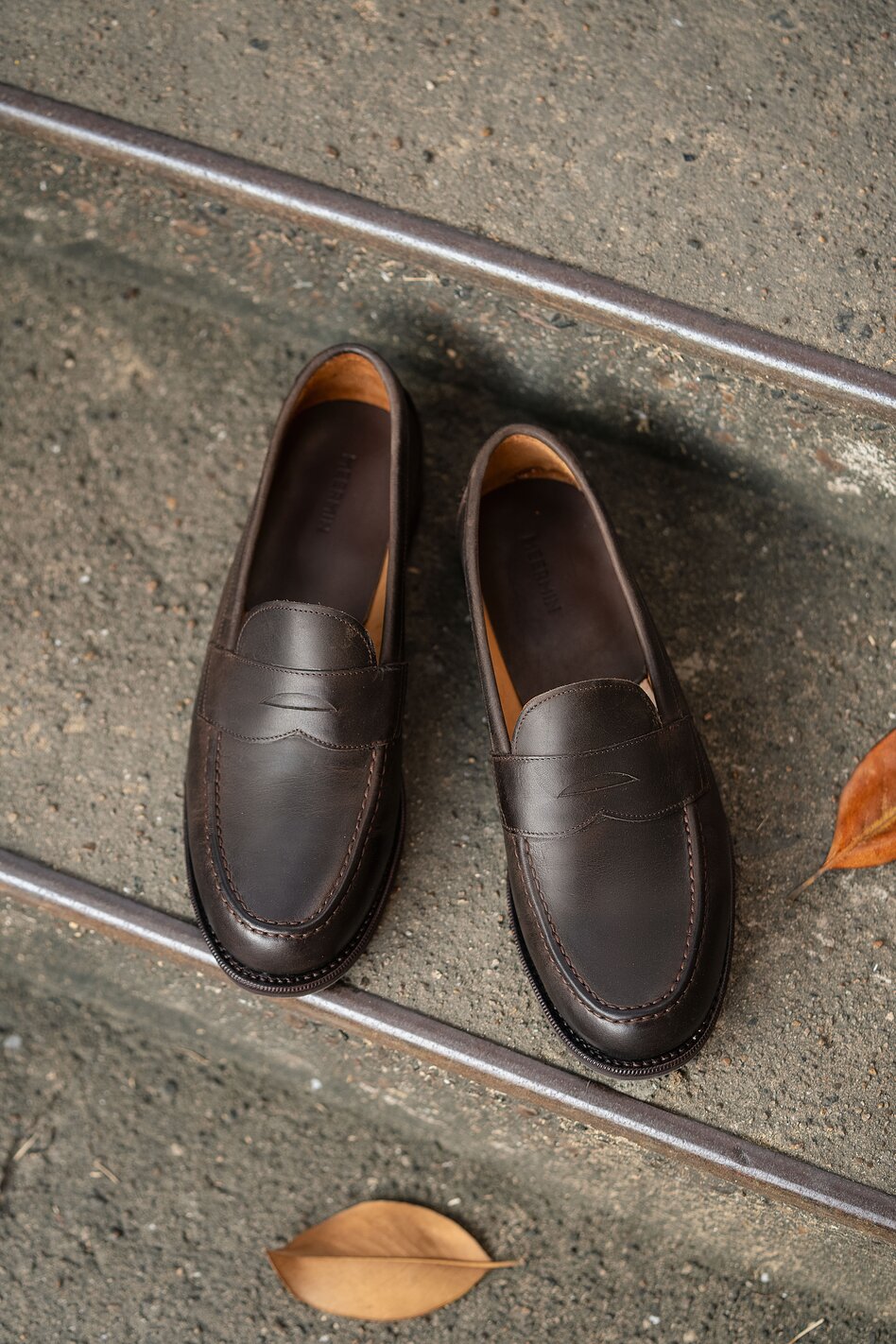 Pair of dark brown leather penny loafers photographed from above on stone steps, showcasing handcrafted construction and timeless design.