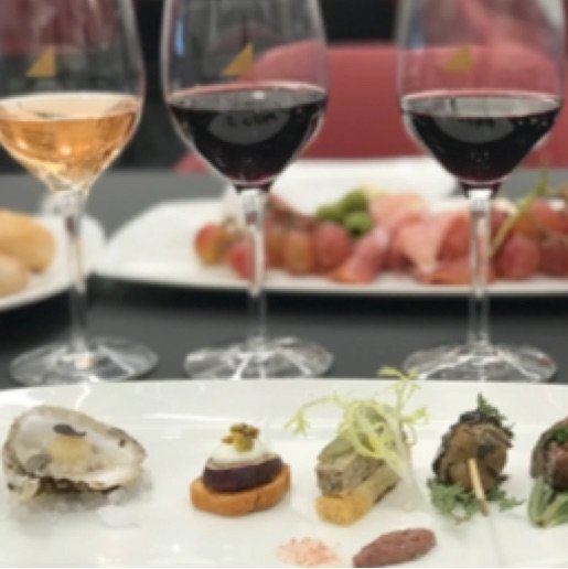 Two glasses of red wine and one white wine stand between two white plates, one embellished with small hors d'oeuvres and one with a red and pink charcuterie. 