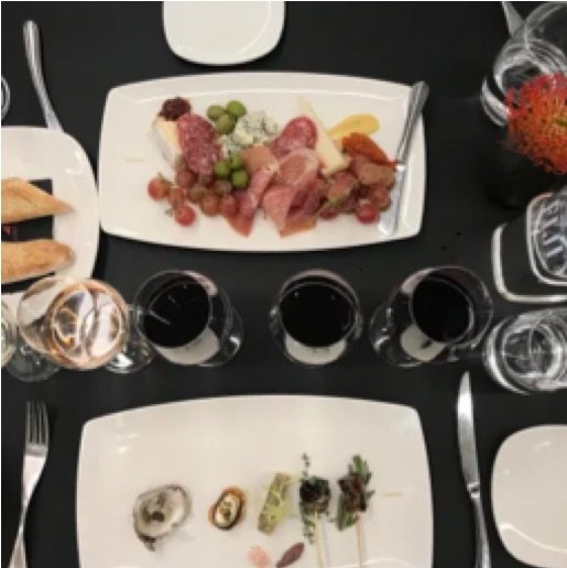  Two glasses of red wine and one white wine stand between two white plates, one embellished with small hors d'oeuvres and one with a red and pink charcuterie. 
