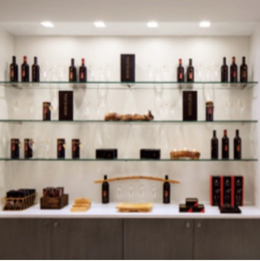 A gift shop wall with glass shelves of wine bottles and accessories, illuminated with gallery style lighting.