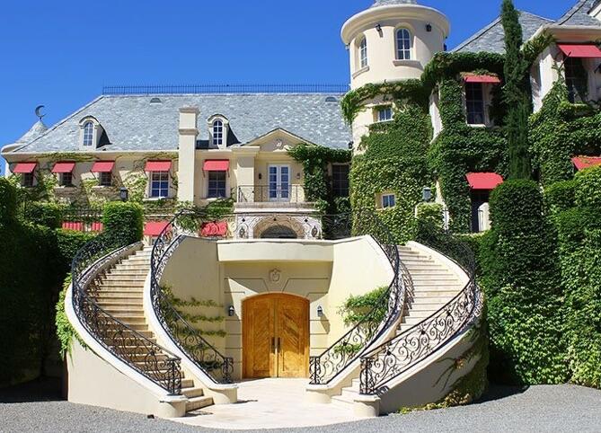 A cream, French-style mansion, coated with ivy, with red awnings and a grand double staircase out front.
