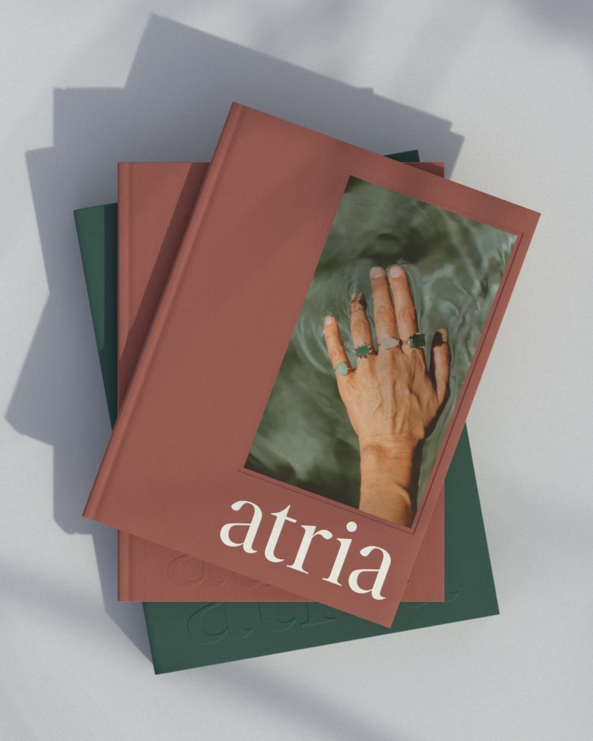A stack of green and red Atria books with a hand in rippling water, on a grey background with shadows to create a calming energy