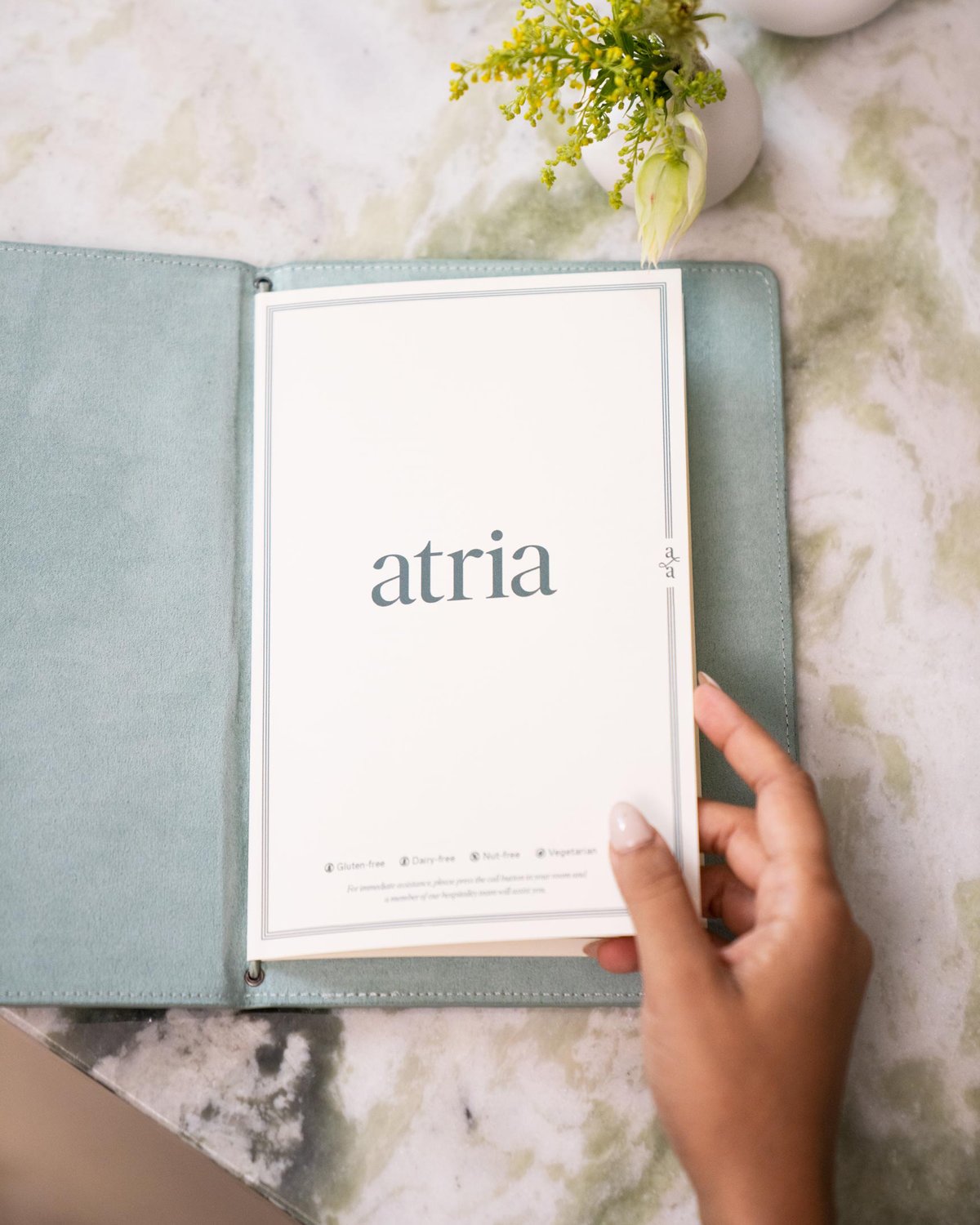 A manicured hand turning the pages of a mint green, designed Atria menu for food and drinks