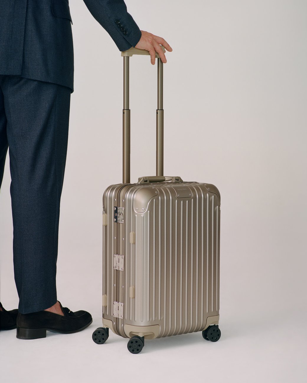 A professional woman in a suit holding a metallic Atria branded gold rolling suitcase with wheels on a white backdrop