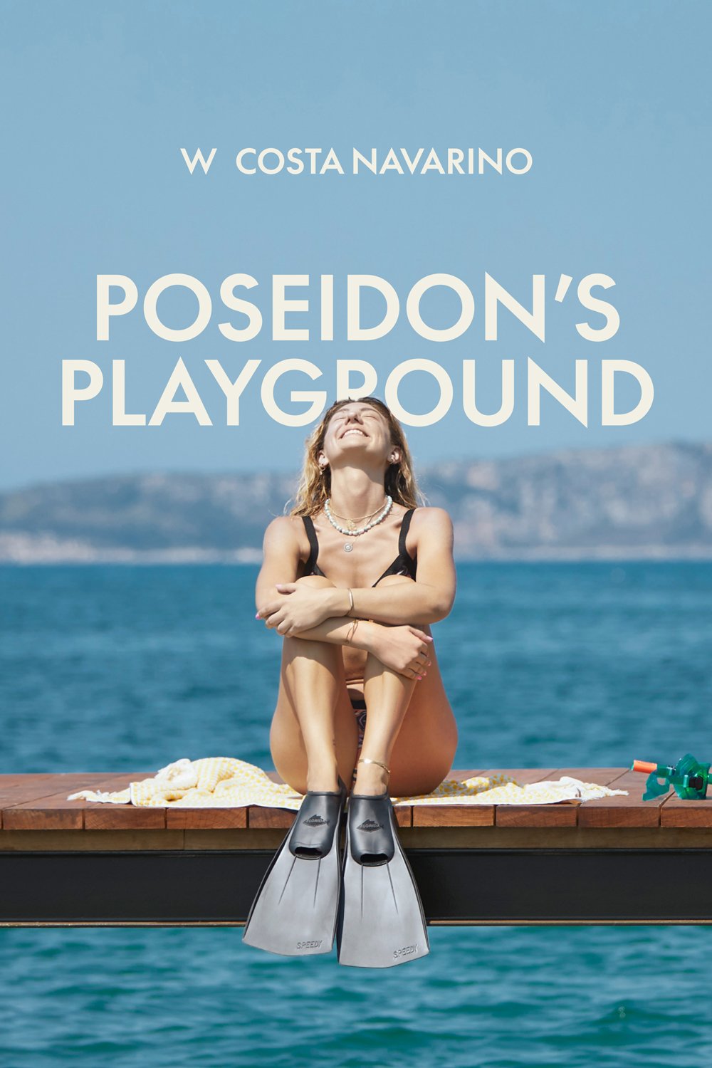 A blonde woman in a bathing suit smiling at the blue sky and blue ocean with flippers on her feet and Greek mountains behind her. The text reads W Hotels Costa Navarino: Poseidon’s Playground in Greece