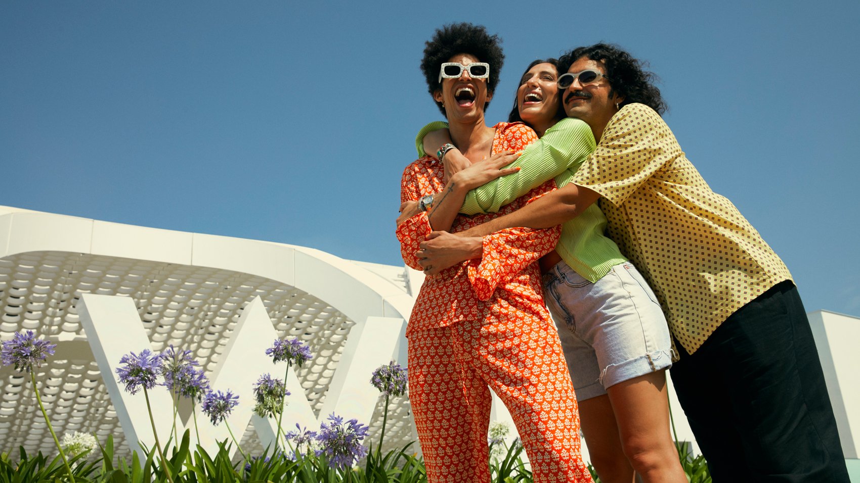 Friends hugging on an international W Hotels vacation in bright clothing with blue skies and happy, smiling faces in front of the hotel sculptural logo