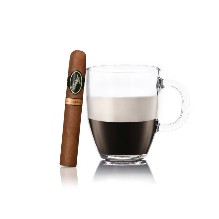 Davidoff Nicaragua Toro cigar beside a frothy Biscuit cocktail in a clear cup on a white background.