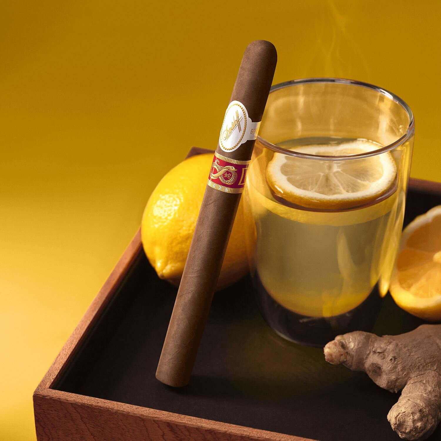 A top-down view of a lemon ginger' tea with a lemon garnish, set on a tray alongside fresh lemons, ginger, and a Lancero Year of the Snake 2025 cigar placed next to the drink.