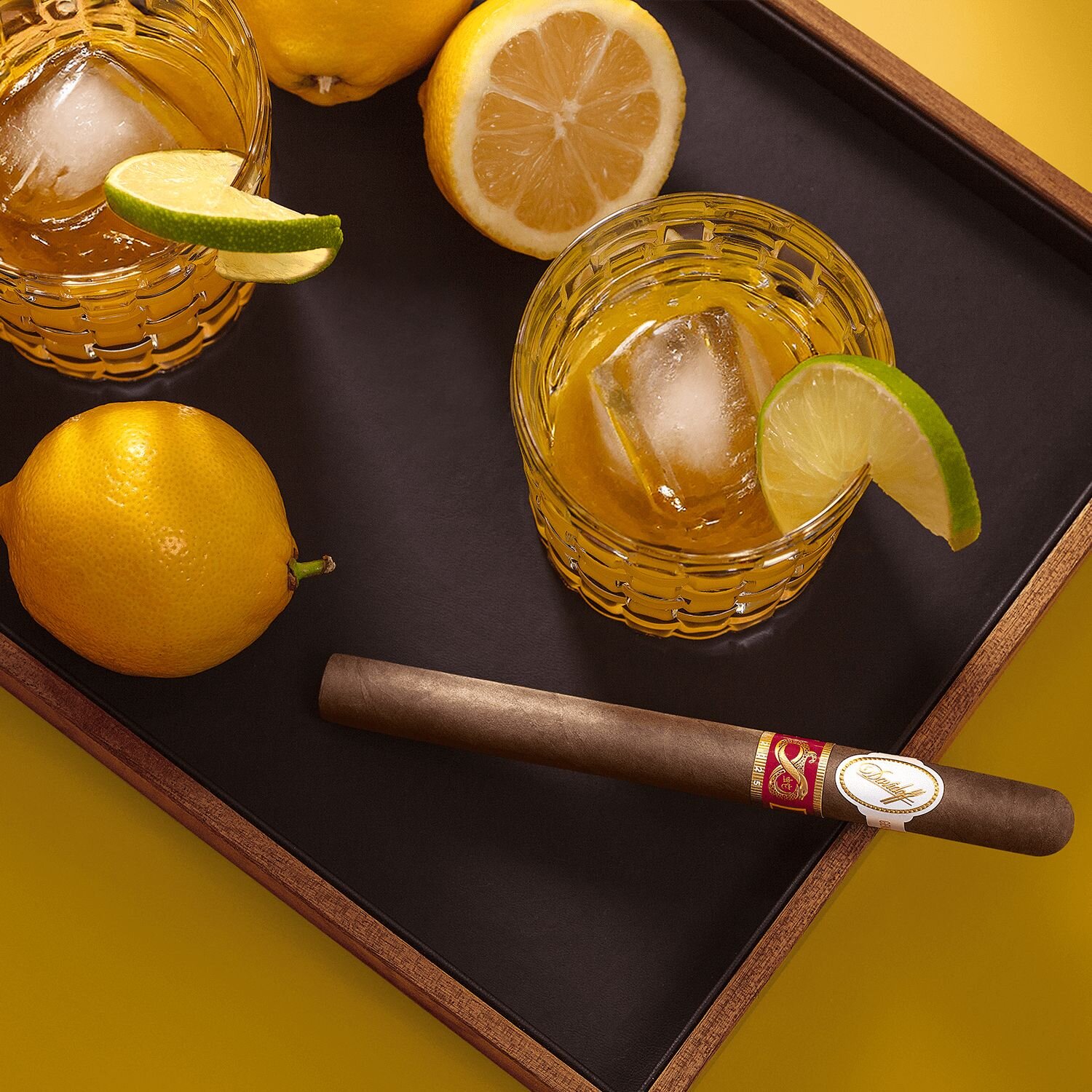 A top-down view of a 'Yellow Bird' cocktail with a lemon garnish, placed on a tray alongside fresh lemons and a Lancero Year of the Snake 2025 cigar.