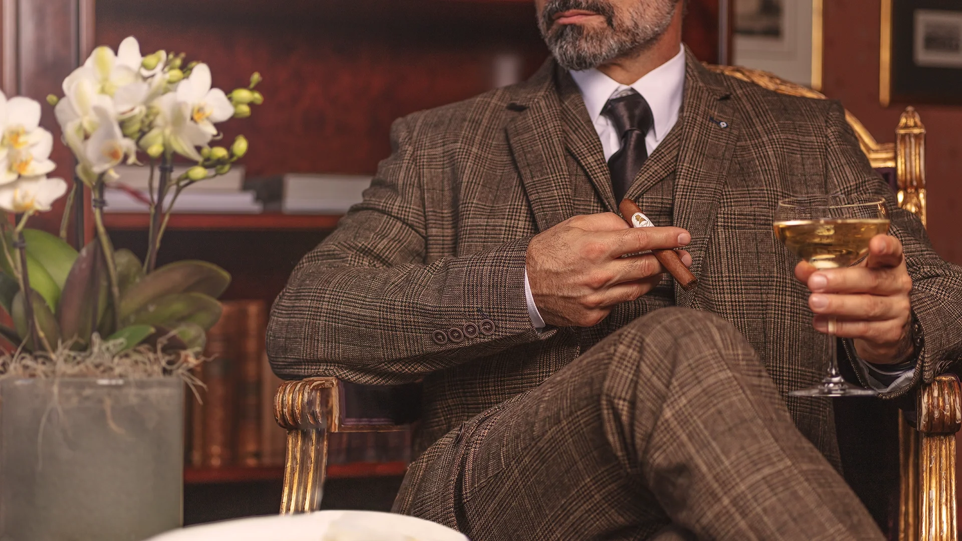 A well-dressed man in a plaid suit sits in a luxurious chair, holding a cigar and a cocktail, surrounded by a sophisticated setting.