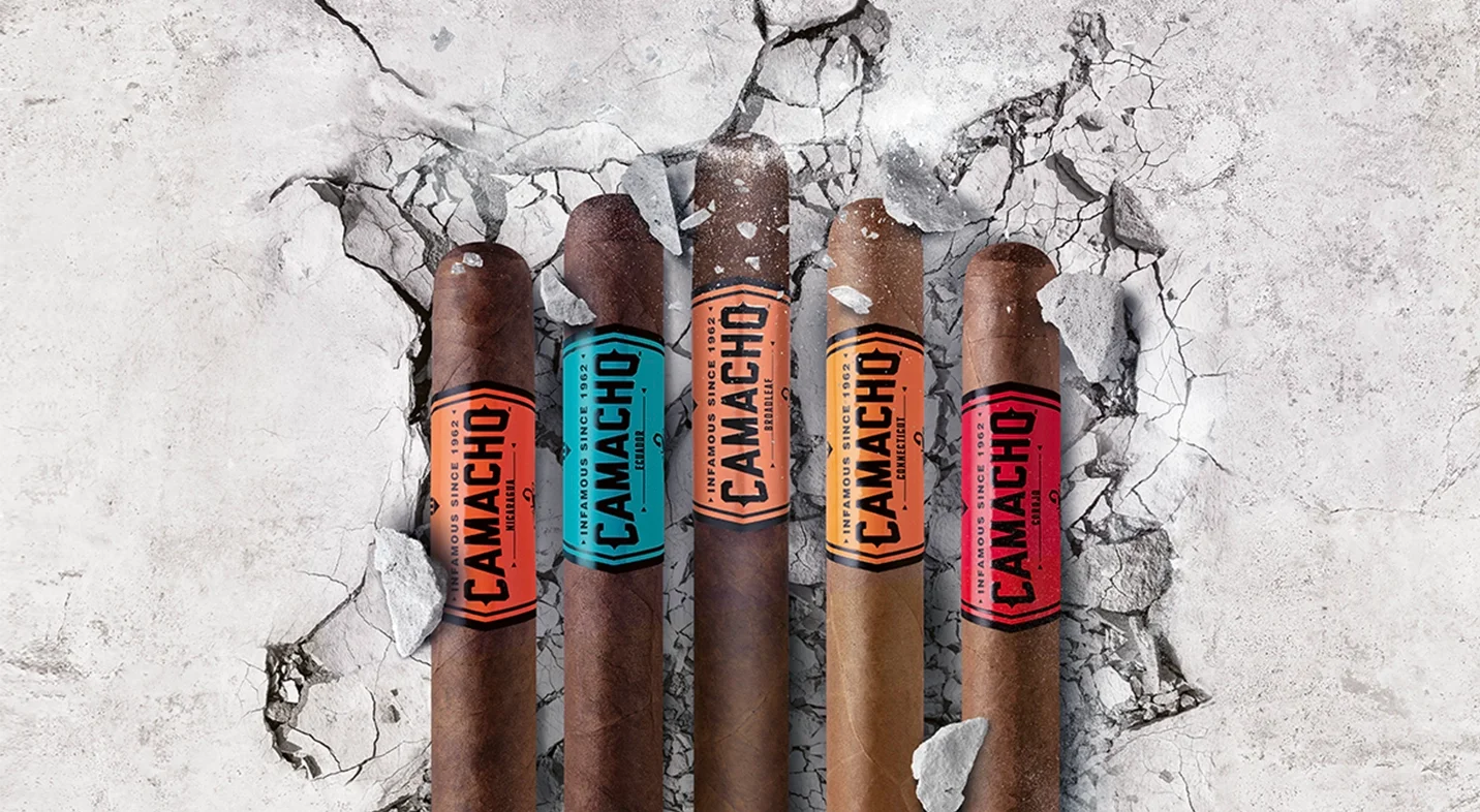 Four Camacho cigars with colorful bands stand against a cracked concrete background, accompanied by the slogan "Built Bold, Rolled for the Rebel."