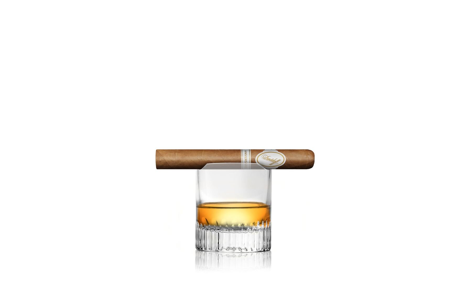 A premium cigar rests atop a whiskey glass filled with amber-colored whiskey, set against a minimal white background.