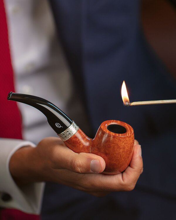 A close-up reveals a hand delicately holding an exquisite pipe, with a lighted match poised above, ready to ignite the tobacco within.
